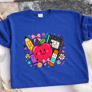 May include: A royal blue t-shirt featuring a colorful graphic print of a smiling red apple, a yellow pencil, a purple crayon, a black and white composition notebook, and an orange glue stick, all decorated with small bows, flowers, and stars.
