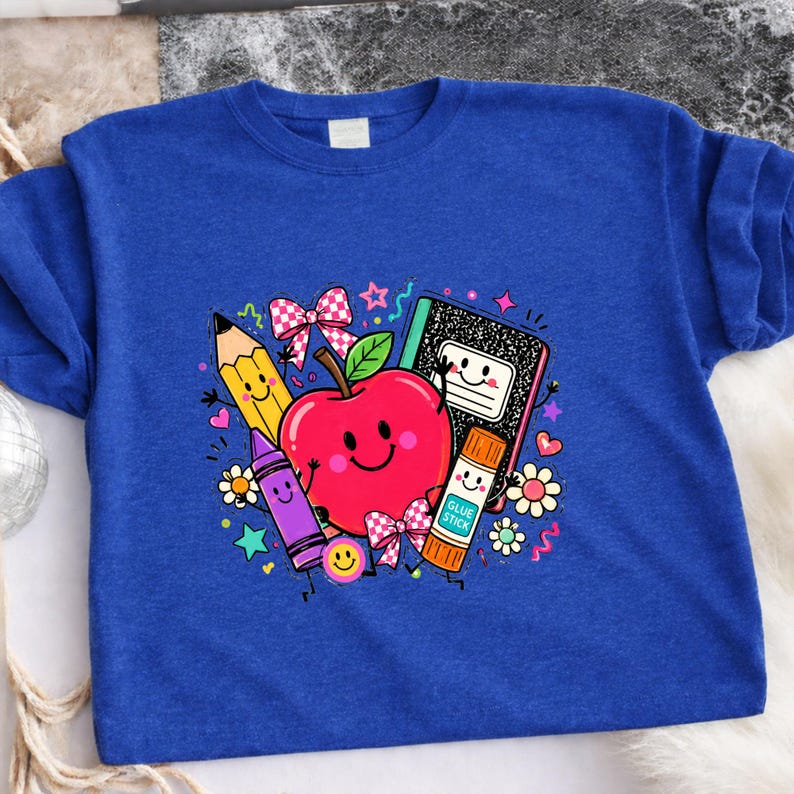 May include: A royal blue t-shirt featuring a colorful graphic print of a smiling red apple, a yellow pencil, a purple crayon, a black and white composition notebook, and an orange glue stick, all decorated with small bows, flowers, and stars.