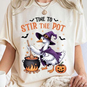 May include: Cream-colored t-shirt with a Halloween-themed graphic. The design features a goose wearing a witch hat and cape, stirring a cauldron. Text above reads "TIME TO STIR THE POT". A jack-o'-lantern and bats complete the design.