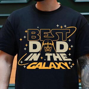 May include: Black t-shirt with a yellow and orange graphic that says "Best Dad in the Galaxy" with a Darth Vader silhouette in the middle.