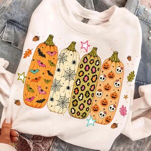 May include: A white crewneck sweatshirt featuring a graphic print of four tall pumpkins. The pumpkins are decorated with colourful bats, spider webs, leopard print, and a pattern of small jack-o'-lanterns and skulls, accented by stars and acorns.