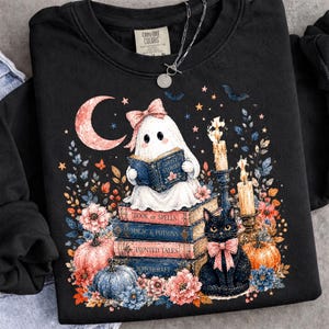 May include: A black crewneck sweatshirt featuring a graphic of a white ghost with a pink bow reading a book titled Ghost Stories, stacked on books labeled Book of Spells, Magic & Potions, Haunted Tales, and Witchcraft, with a black cat and pumpkins.