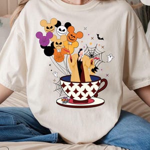Pu&ograve; includere: T-shirt color crema con un design stravagante di Halloween. La grafica include un personaggio in una tazza da t&egrave;, circondato da palloncini a forma di teste di Topolino, ragnatele, fantasmi e pipistrelli. I palloncini sono di vari colori.