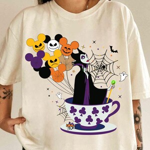Pu&ograve; includere: T-shirt color crema con un design a tema Halloween. La grafica include un personaggio di strega in una tazza da t&egrave;, circondato da palloncini a forma di teste di Topolino con espressioni spettrali, una ragnatela e fantasmi.