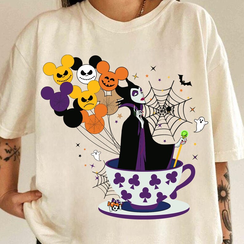 Pu&ograve; includere: T-shirt color crema con un design a tema Halloween. La grafica include un personaggio di strega in una tazza da t&egrave;, circondato da palloncini a forma di teste di Topolino con espressioni spettrali, una ragnatela e fantasmi.