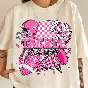 May include: Cream-coloured t-shirt featuring a pink football helmet, football, and megaphone design. The words "TACKLE Cancer CHEER" are displayed in pink. The football and helmet have pink ribbon accents.