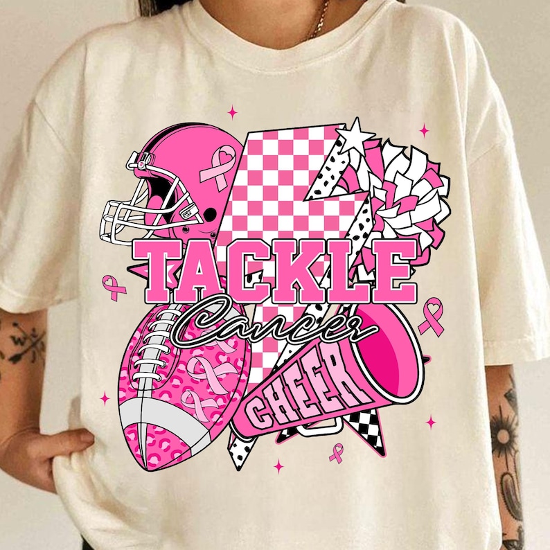 May include: Cream-coloured t-shirt featuring a pink football helmet, football, and megaphone design. The words "TACKLE Cancer CHEER" are displayed in pink. The football and helmet have pink ribbon accents.