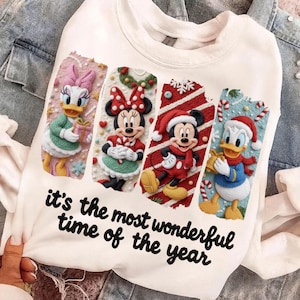 Könnte beinhalten: Weißes Sweatshirt mit vier Disney-Figuren: Daisy Duck, Minnie Mouse, Mickey Mouse und Donald Duck, alle in festlicher Kleidung. Der Text "it's the most wonderful time of the year" ist unter den Figuren aufgedruckt.