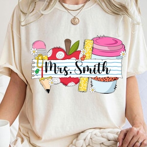K&ouml;nnte beinhalten: Cremefarbenes T-Shirt mit farbenfrohem Grafikdesign. Das Design umfasst einen Apfel, einen Bleistift, ein Lineal und einen Beh&auml;lter mit dem Text "Mrs. Smith" auf einem linierten Papierbanner. Das Design ist mit kleinen wei&szlig;en Blumen verziert.
