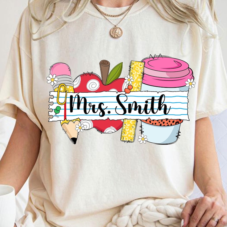 K&ouml;nnte beinhalten: Cremefarbenes T-Shirt mit farbenfrohem Grafikdesign. Das Design umfasst einen Apfel, einen Bleistift, ein Lineal und einen Beh&auml;lter mit dem Text "Mrs. Smith" auf einem linierten Papierbanner. Das Design ist mit kleinen wei&szlig;en Blumen verziert.