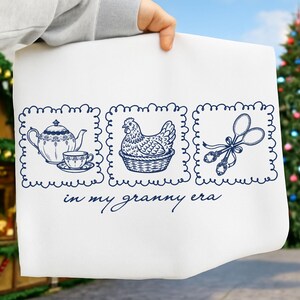 May include: White tea towel with a navy blue illustration featuring a teapot and teacup, a hen in a basket, and kitchen utensils. The text "in my granny era" is written below the illustrations.