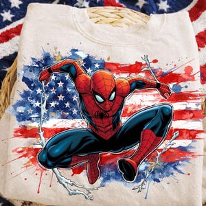 Puede incluir: Sudadera beige con un gráfico dinámico de Spider-Man. El superhéroe está representado en su traje clásico rojo y azul, sobre un fondo de la bandera estadounidense con salpicaduras de pintura roja, blanca y azul.