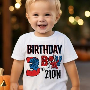 May include: White t-shirt with a red, blue, and black Spiderman graphic. The shirt says "Birthday Boy" and "3" with a spider web design. The name "Zion" is written below the number.