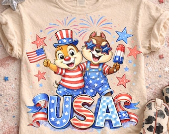 Può includere: T-shirt beige con un design patriottico. Due scoiattoli dei cartoni animati indossano occhiali da sole e tengono in mano una bandiera americana e un ghiacciolo. La parola "USA" è visualizzata in rosso, bianco e blu con accenti di stelle. Fuochi d'artificio e stelle completano il design.