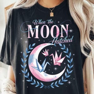 May include: Black t-shirt with a graphic of a crescent moon with pink and blue watercolor, three origami cranes, and the text "When the Moon Hatched".