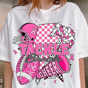 May include: White t-shirt featuring a pink American football helmet, ball, and megaphone graphic. The text reads "TACKLE Cancer CHEER". The ball has a pink ribbon pattern. Pink ribbons and stars are scattered around the design.