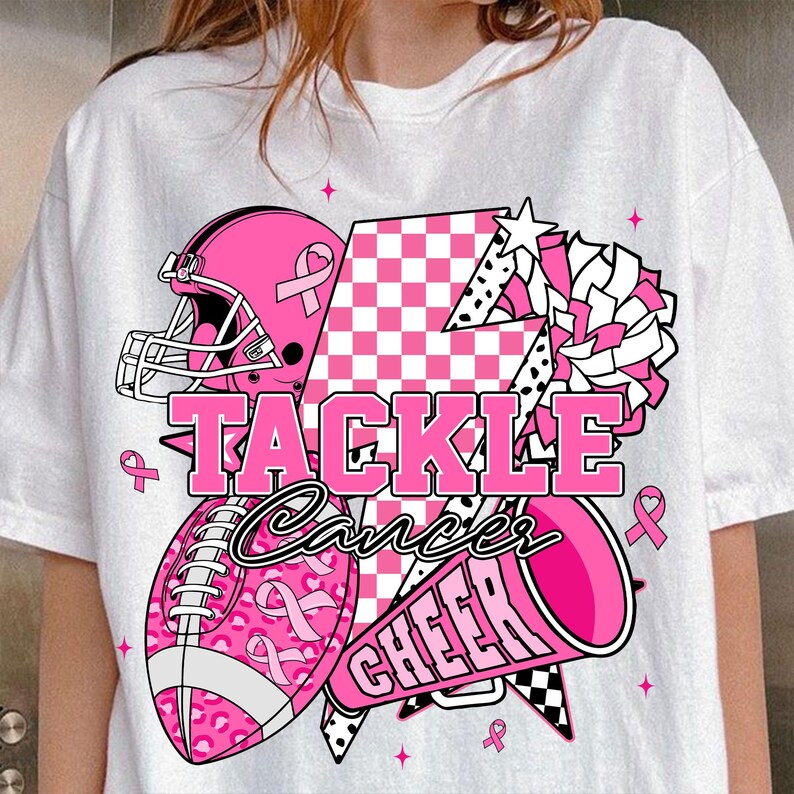 May include: White t-shirt featuring a pink American football helmet, ball, and megaphone graphic. The text reads "TACKLE Cancer CHEER". The ball has a pink ribbon pattern. Pink ribbons and stars are scattered around the design.