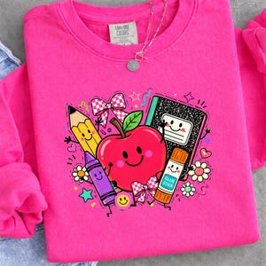 May include: A bright pink crewneck sweatshirt featuring a colorful graphic of a smiling red apple, a yellow pencil, a purple crayon, a black and white composition notebook, and a glue stick labeled GLUE STICK, accented with bows and stars.