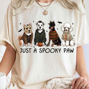 Op de afbeelding: Crèmekleurig T-shirt met de tekst "JUST A SPOOKY PAW". De afbeelding toont vier honden verkleed als horrorfilmkarakters, waaronder een hond met een hockeymasker, een hond in een groene jas en een hond in een gestreepte trui.
