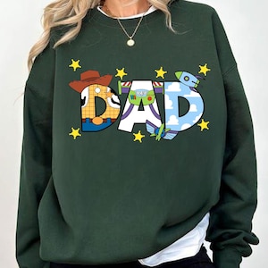 May include: A dark green sweatshirt with a graphic print of the word "DAD" made up of cartoon characters from the Toy Story movies. The characters include Woody, Buzz Lightyear, and a green alien. The graphic is surrounded by yellow stars.
