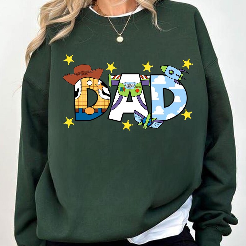 May include: A dark green sweatshirt with a graphic print of the word "DAD" made up of cartoon characters from the Toy Story movies. The characters include Woody, Buzz Lightyear, and a green alien. The graphic is surrounded by yellow stars.