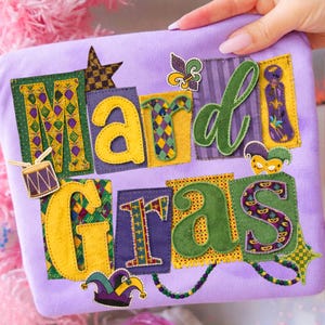 May include: A purple fabric item with the words "Mardi Gras" appliquéd in various colors. The letters are yellow, green, and purple, with decorative elements like a star, fleur-de-lis, and a jester's mask. A drum is also included.