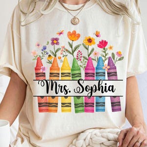 May include: Cream-colored t-shirt featuring a colorful crayon design with flowers and butterflies. The text "Mrs. Sophia" is written in cursive across the crayons. The crayons are red, orange, yellow, green, pink, blue, and purple.