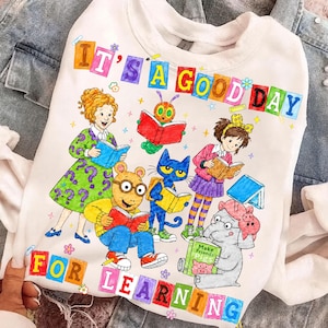 May include: A white crewneck sweatshirt featuring a colorful graphic of various storybook characters reading books. The text "IT'S A GOOD DAY FOR LEARNING" is printed in block letters above and below the characters, surrounded by small stars.