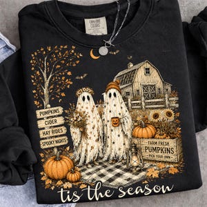 May include: A black crewneck sweatshirt featuring a graphic of two ghosts, one holding a bouquet and the other a pumpkin mug. The design includes a barn, pumpkins, a lantern, a sign reading PUMPKINS CIDER HAY RIDES SPOOKY NIGHTS, and the text tis the season.
