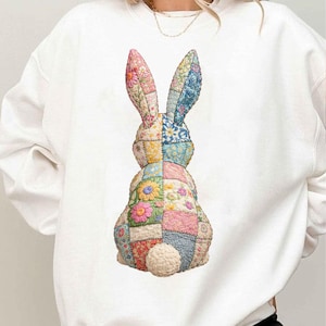 Könnte beinhalten: Weißes Sweatshirt mit einem farbenfrohen Patchwork-Hasen-Design. Der Hase hat große Ohren und einen flauschigen Schwanz. Der Patchwork-Stoff enthält Blumen- und geometrische Muster in Rosa, Blau, Gelb und Grün.