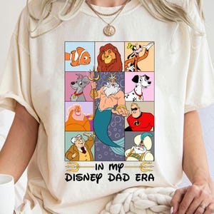 May include: A white t-shirt with a graphic of various Disney characters in a grid pattern. The characters include Ariel, Nemo,  Goofy,  and more. The text "In My Disney Dad Era" is printed below the graphic.