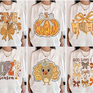 May include: A collection of six white t-shirts with fall-themed graphics. Designs include the phrases "Give Thanks," "Cozy Season," and "God Says I Am," along with images of pumpkins, bows, and a turkey. The shirts feature orange, brown, and yellow colors.