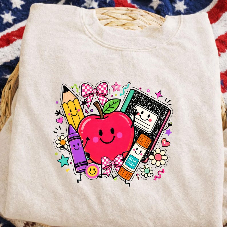 May include: A cream-colored crewneck sweatshirt featuring a colorful graphic print of a smiling red apple, a yellow pencil, a purple crayon, a black and white composition notebook, and a glue stick, all decorated with pink bows and small flowers.