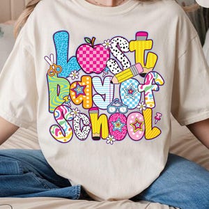 Może przedstawiać: Biały T-shirt z kolorową grafiką, na kt&oacute;rej widnieje napis "Ostatni dzień szkoły". Grafika składa się z r&oacute;żnych kształt&oacute;w i wzor&oacute;w, w tym gwiazd, kropek, pask&oacute;w i wzor&oacute;w w kratkę.