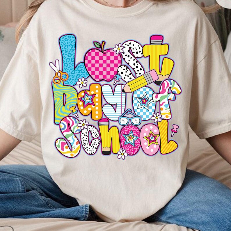 Może przedstawiać: Biały T-shirt z kolorową grafiką, na kt&oacute;rej widnieje napis "Ostatni dzień szkoły". Grafika składa się z r&oacute;żnych kształt&oacute;w i wzor&oacute;w, w tym gwiazd, kropek, pask&oacute;w i wzor&oacute;w w kratkę.