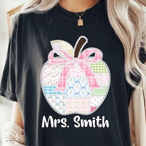 May include: Black t-shirt featuring a patchwork apple design with a pink bow and lace trim. The apple is filled with various patterns and colors. The name "Mrs. Smith" is printed in white below the apple.
