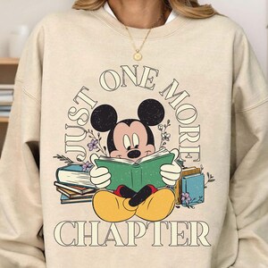 Op de afbeelding: Een beige sweatshirt met een afbeelding van Mickey Mouse die een boek leest. De tekst rond Mickey Mouse zegt "Just One More Chapter".