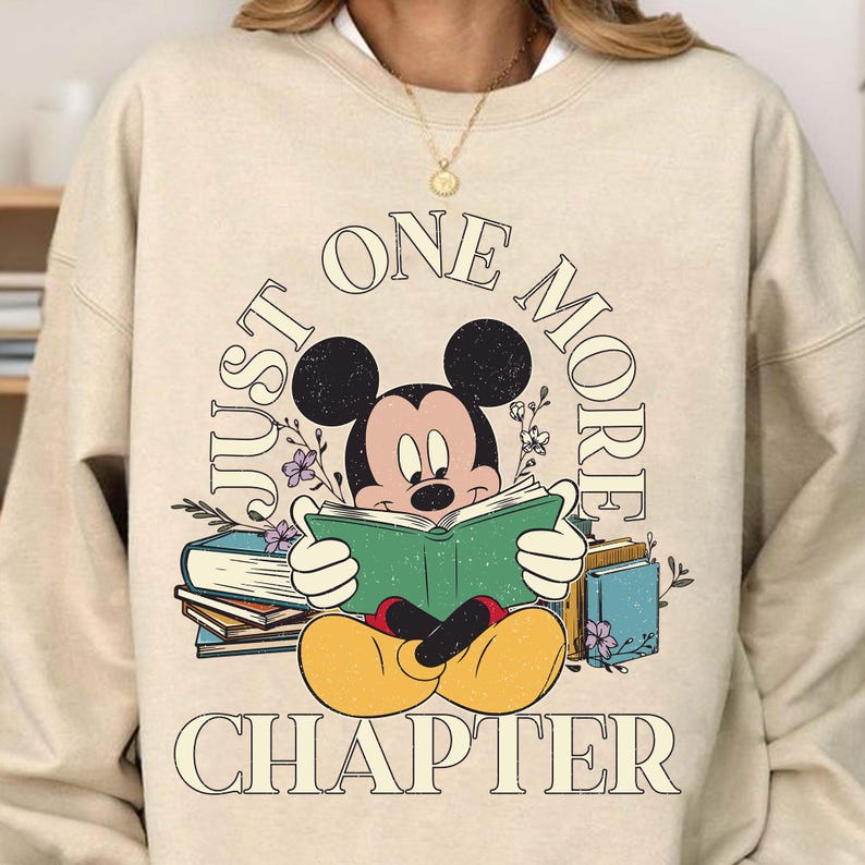 Op de afbeelding: Een beige sweatshirt met een afbeelding van Mickey Mouse die een boek leest. De tekst rond Mickey Mouse zegt "Just One More Chapter".