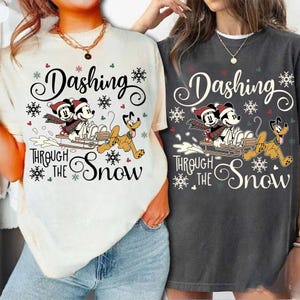 May include: Two t-shirts, one cream and one charcoal, feature a festive design. The graphic includes Mickey and Minnie Mouse on a sled with Pluto, snowflakes, and the text "Dashing Through The Snow".
