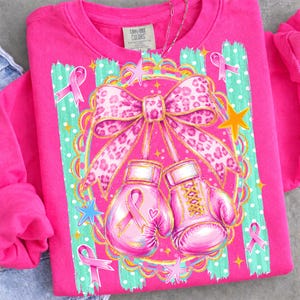 May include: A bright pink long-sleeve sweatshirt featuring a graphic of two pink boxing gloves tied together with a large leopard-print bow. The design includes pink awareness ribbons, yellow stars, and a teal striped background with white dots.