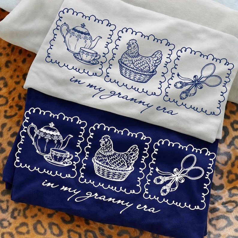May include: Two t-shirts, one cream and one navy blue, with a whimsical design. Each shirt features three framed illustrations: a teapot and teacup, a hen in a basket, and a pair of spoons. The text "in my granny era" is below the illustrations.