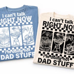 May include: Two t-shirts, one blue and one cream, with the text "i can't talk RIGHT NOW" above three illustrated panels. The panels depict a cooler with fish, a grill, and a lawnmower. Below is the text "i'm doing hot DAD STUFF".