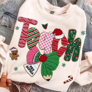 May include: Cream-colored sweatshirt with the word "Teach" in a festive, textured design. The letters are adorned with Christmas-themed patterns, including candy canes, gingerbread men, and a Santa hat. A bow and pencil design are also featured.