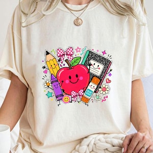 May include: A cream-colored t-shirt featuring a colorful graphic print of a smiling red apple surrounded by school supplies including a yellow pencil, a purple crayon, a black and white composition notebook, and a glue stick with a pink bow.