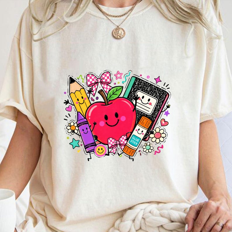 May include: A cream-colored t-shirt featuring a colorful graphic print of a smiling red apple surrounded by school supplies including a yellow pencil, a purple crayon, a black and white composition notebook, and a glue stick with a pink bow.