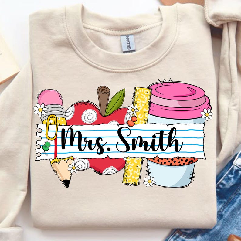 K&ouml;nnte beinhalten: Beiges Sweatshirt mit einem Lehrer-Themen-Design. Das Design umfasst einen roten Apfel, einen Bleistift, ein Lineal und eine Kaffeetasse mit dem Namen "Mrs. Smith" auf einem linierten Papierbanner. Kleine G&auml;nsebl&uuml;mchen-Akzente.