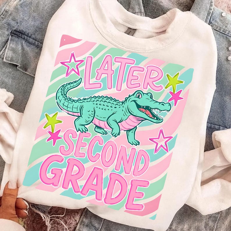 Peut inclure: Sweat-shirt blanc avec une illustration d'alligator vert d'eau et le texte "LATER SECOND GRADE" en rose. Le motif comprend des &eacute;toiles roses et vertes d'eau et un fond ray&eacute; aux couleurs pastel.