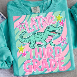 Peut inclure: Sweat-shirt turquoise avec un motif d'alligator vert, d'&eacute;toiles roses et les mots "LATER THIRD GRADE" en rose. Le motif est plac&eacute; sur un fond ray&eacute; rose et vert.