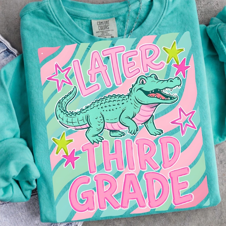 Peut inclure: Sweat-shirt turquoise avec un motif d'alligator vert, d'&eacute;toiles roses et les mots "LATER THIRD GRADE" en rose. Le motif est plac&eacute; sur un fond ray&eacute; rose et vert.
