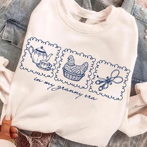 May include: White sweatshirt with a blue graphic design featuring a teapot and teacup, a hen in a basket, and kitchen scissors. The phrase "in my granny era" is written below the graphics.